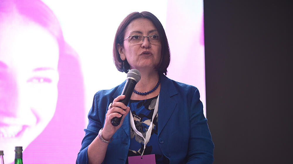 A woman in a blue blazer holds a microphone and speaks