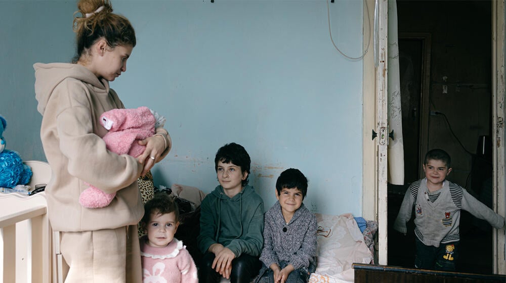 A woman is standing holding a baby. Four young children are also in the room. They are all smiling.