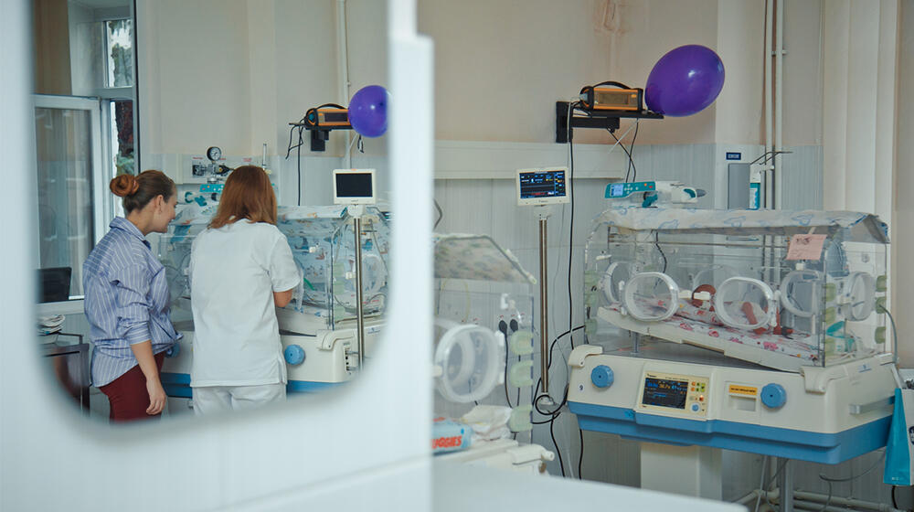 A maternity ward seen through an opened door. Three incubators are visible and a nurse & a woman with their backs to the camera.