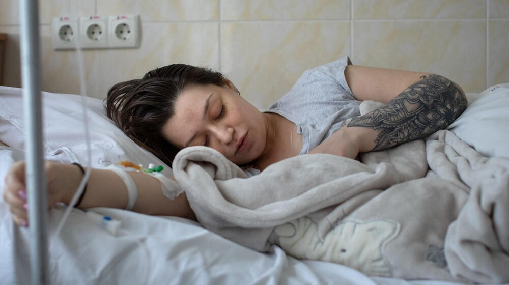 A woman lays in a hospital bed with her eyes closed. Next to her is a blanket wrapped up around a baby.