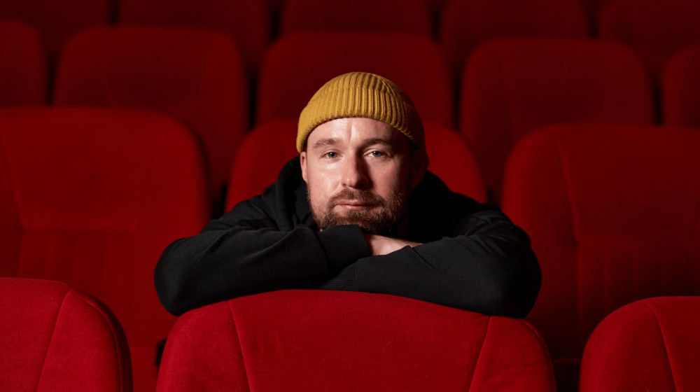 Bohdan Zhuk in a yellow beanie sits in a red movie theatre seat, leaning forward with his arms crossed over the top of the seat