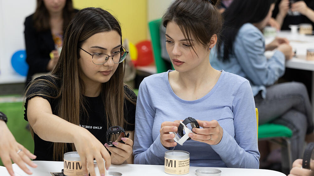 Two young women sit at a desk in a classroom, looking at watches