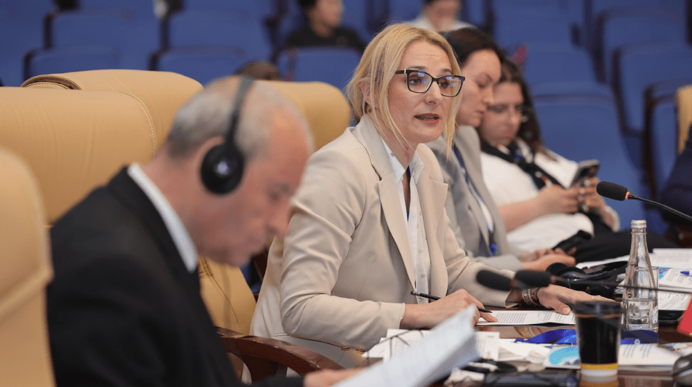 Woman with blonde hair and glasses speaks at conference while man with headphones listens in foreground