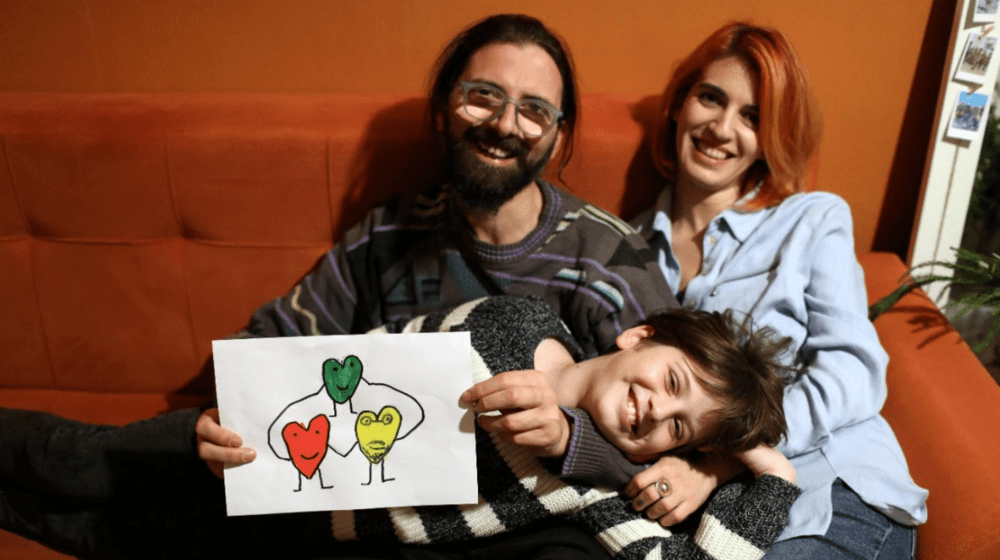 A man, woman and child are sitting together. The child is holding a piece of artwork and there is an orange background behind th