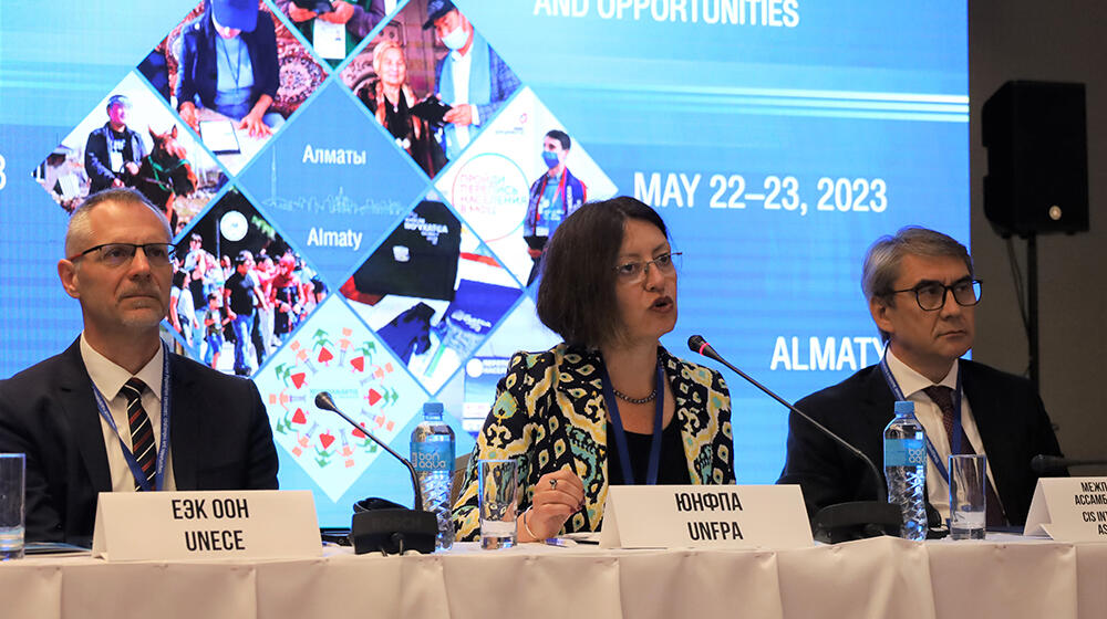 Photo of two men and one woman sitting at a table. Conference background is behind them.