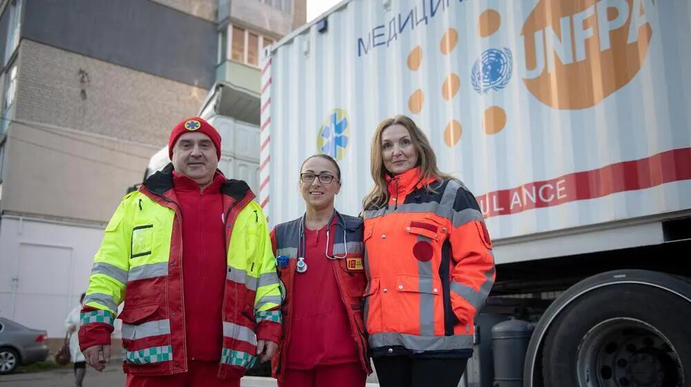 A man and two women, wearing medical clothes, stand outside in front of a truck. They are looking into the camera.