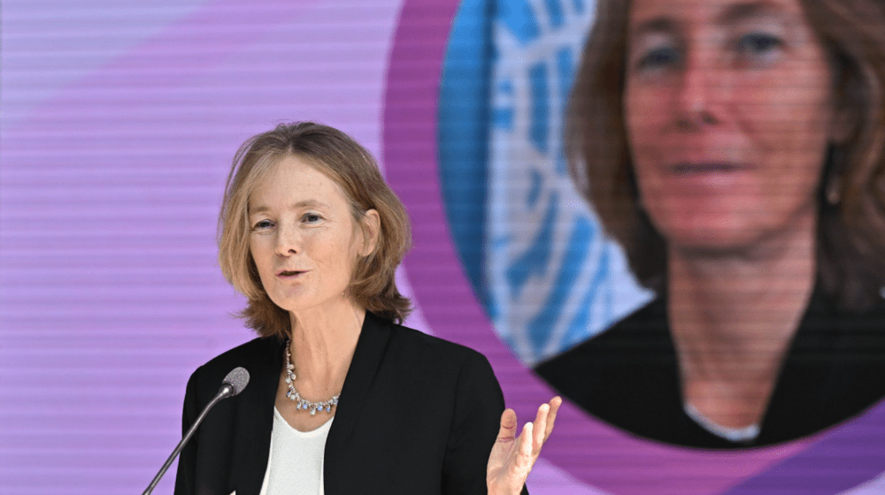 A woman with blonde hair, wearing a black jacket and a white blouse, is standing at podium speaking.