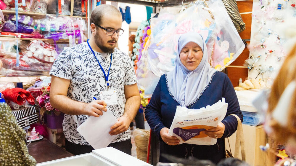 A woman is talking to a clothes seller in his shop. He is wearing glasses. She is wearing a head covering.