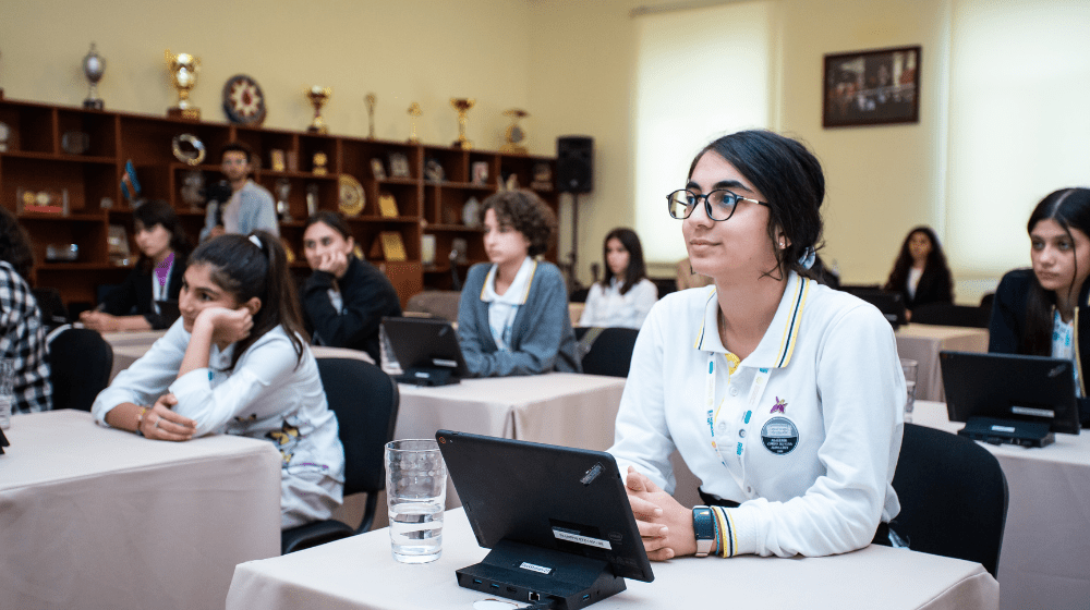 Young girl with dark hair and glasses sits at in a classroom with a tablet on her desk. She is surrounded by other young girls