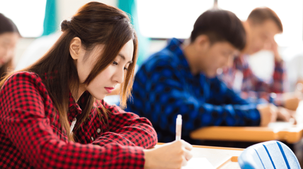 A young woman with dark hair writing at a school desk