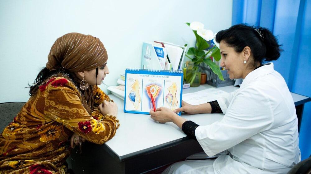 A doctor is talking to a woman in a doctor's room. They are both sitting down.