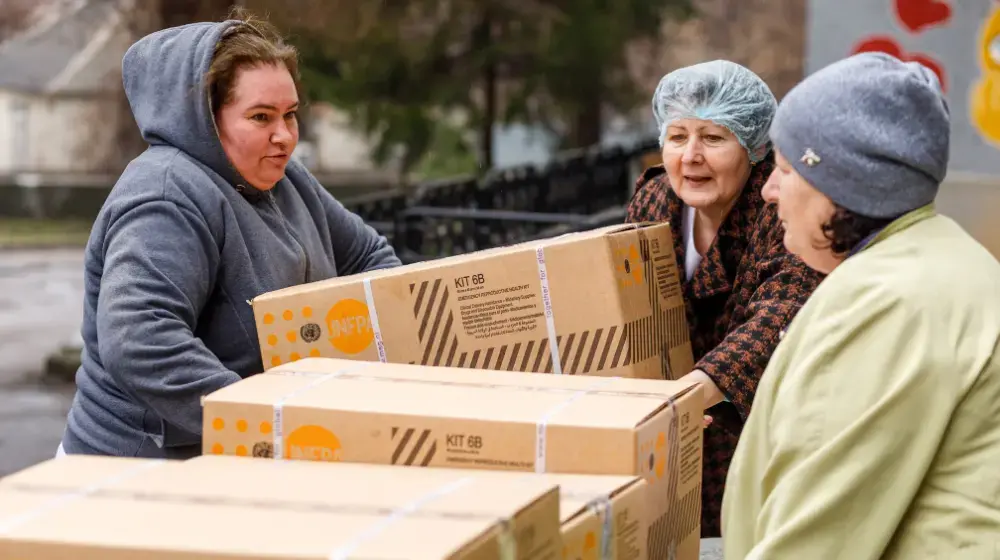 Three women picking up a large cardboard box from a row of similar boxes.