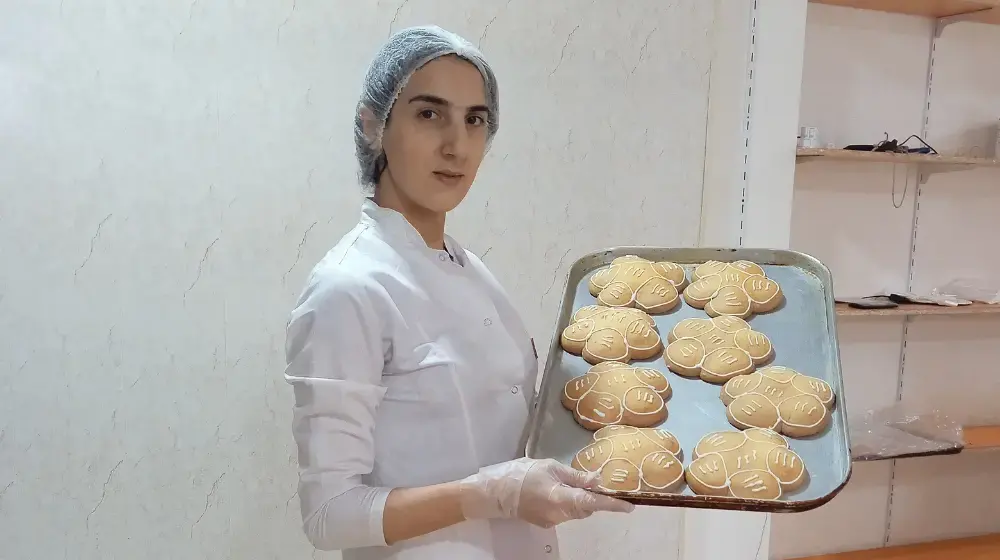 Young woman wearing white chef's jacket, hair net and plastic gloves holds up sheet pan filled with flower-shaped backed goods with white decorative icing on top