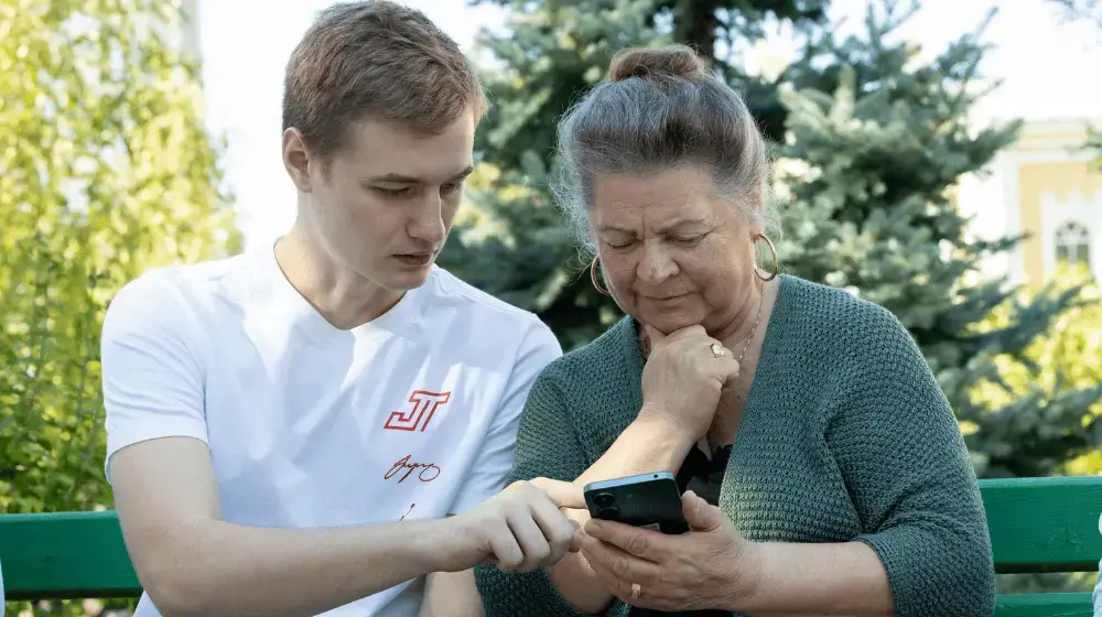 Two people sitting on a wooden bench in an outdoor park. On the right is an older woman with brown hair and grey streaks pulled into a bun wearing a green knit cardigan. She is holding her smartphone in her left hand while gripping her chin with her right hand and staring intently at the screen. On her left, a young man in a white tshirt and light brown hair points to her phone screen as though explaining something to her.