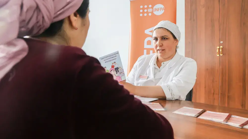 Two women sit at a wooden table inside a room. The camera looks over the right shoulder of one woman wearing a dark maroon sweater and a light purple headscarf. Across from her a woman in white doctor's uniform and cap speaks to her while holding an information brochure. An orange UNFPA banner is in the background