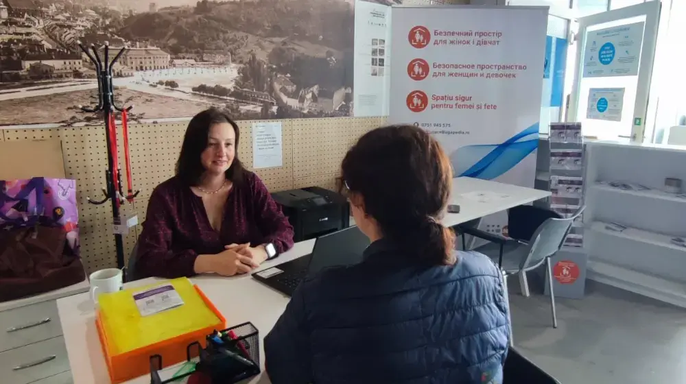 Two women sit at a desk facing each other inside the Safe Space office lobby, one woman with dark brown hair in a low ponytail and black jackets has her back to the camera. The woman across from her greets her with a welcoming smile. A UNFPA Safe Space banner stands in the background.