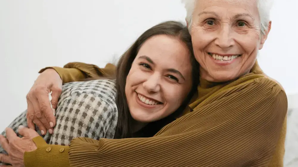 Close up of an older woman with short white hair and a caramel-coloured blouse smiling and wrapping her arms around a younger woman who is also smiling widely at the camera, leaning into the older woman's hug. The young woman has long brown hair and is wearing a green-and-white checked top