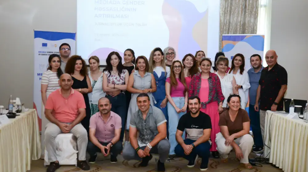 Large group photo of dozens of journalists, bloggers and editors smiling at the camera, mostly standing but some sitting and kneeling in the front row, between long meeting room tables and in front of a projector screen showing a media training presentation. 