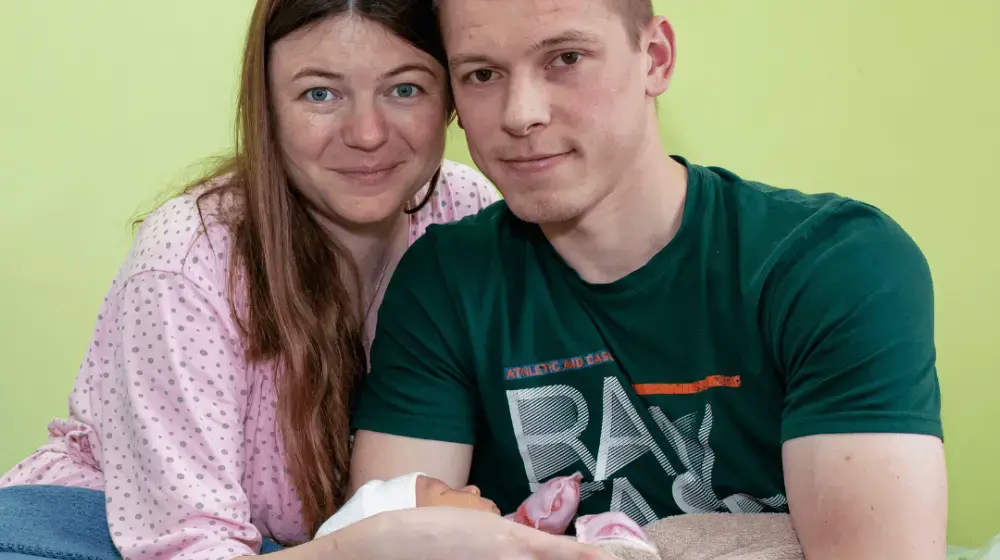 Close up photo of a young couple holding their newborn baby: On the left a woman with long auburn hair in a pink blouse with grey polkadots leans close to a young man in a forest green tshirt with text on the front. They are both smiling lightly at the camera while the man cradles in his arms a newborn baby with a white toque and pink mittens. The background is a solid light green wall.