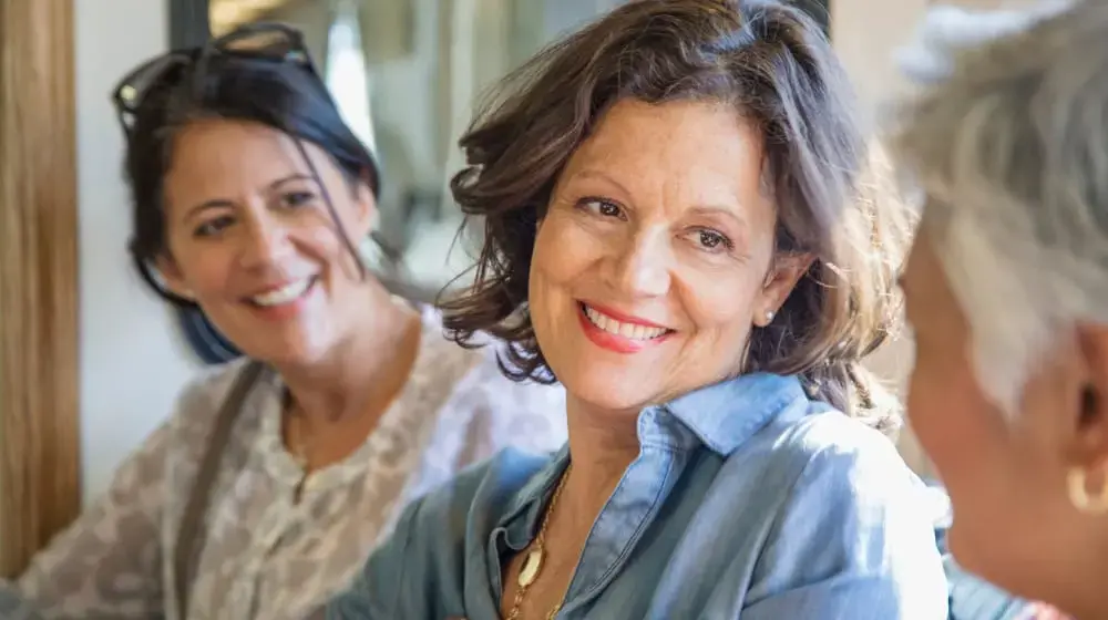 Tight shot of a middle-aged woman with wavy shoulder-length dark brown hair, wearing a losse denim button down shirt. She's smiling and leaning over her left shoulder, toward the camera, talking to a woman with short white hair whose facing away from the camera, visible only from the back of her head. In the background is another woman with dark brown hair pulled into a loose ponytail, wearing a light grey cardigan, smiling and leaning toward the others as though listening in on the conversation.