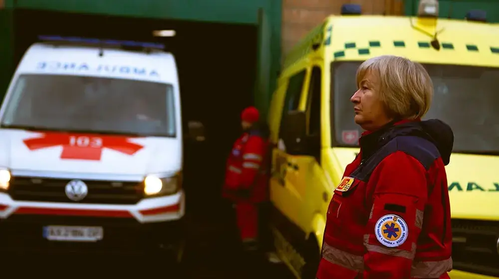 A woman with short blonde hair wearing a red medical jacket stands in front of ambulances