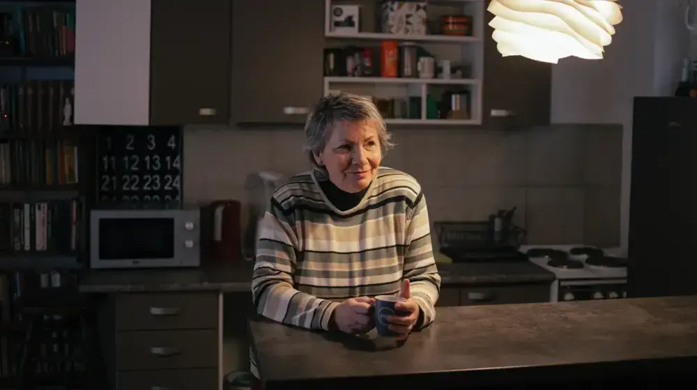 An older woman is sitting at a table in a kitchen. She has her hands around a drinking cup. She is smiling.