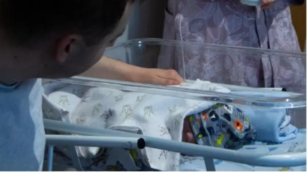 Newborn baby wrapped in blanket and colourful hat lays in hospital crib as father in foreground gently touches the baby