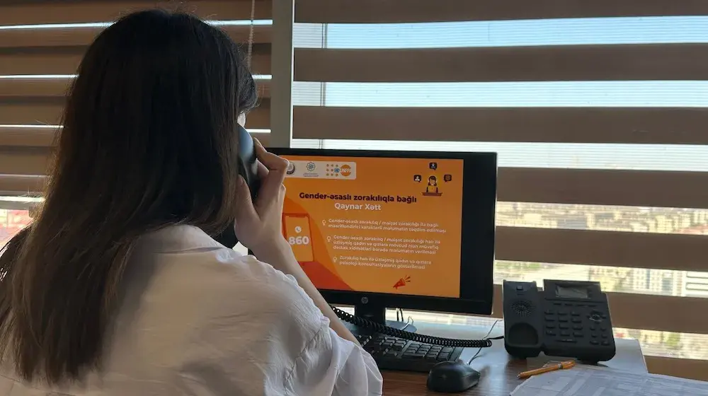 Woman with long brown hair seen from behind as she holds a telephone and looks at a computer screen with the helpline info