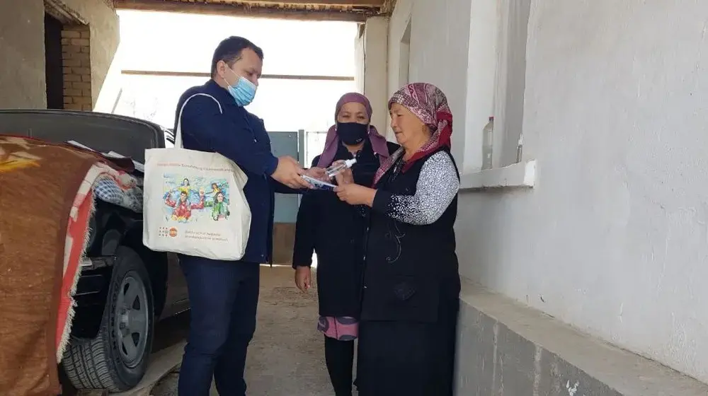 A man is holding a tablet for a woman to sign. Another woman is standing by watching. They are in a garage with a car.