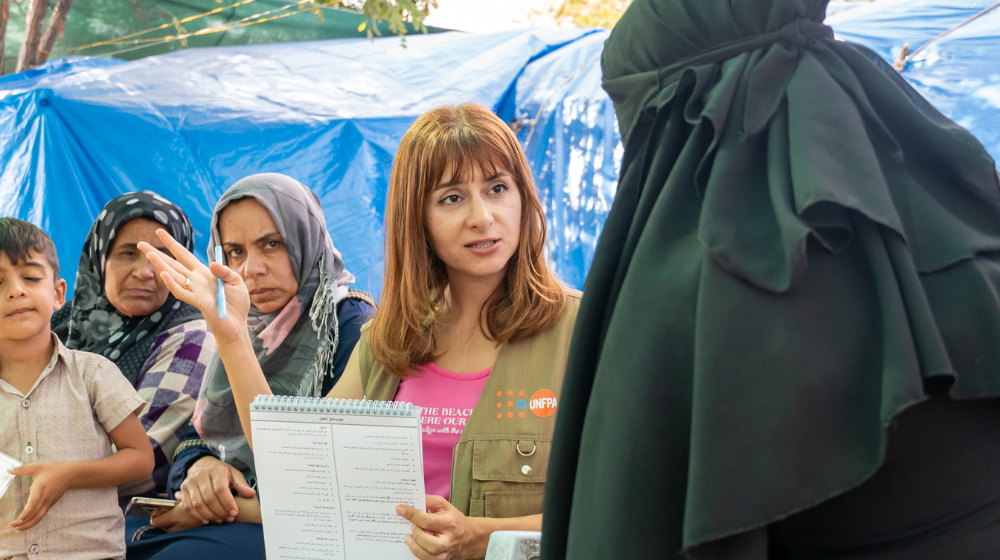 Woman with long brown hair wearing a UNFPA-branded vest and holding a notebook is talking to a woman in a black veil.