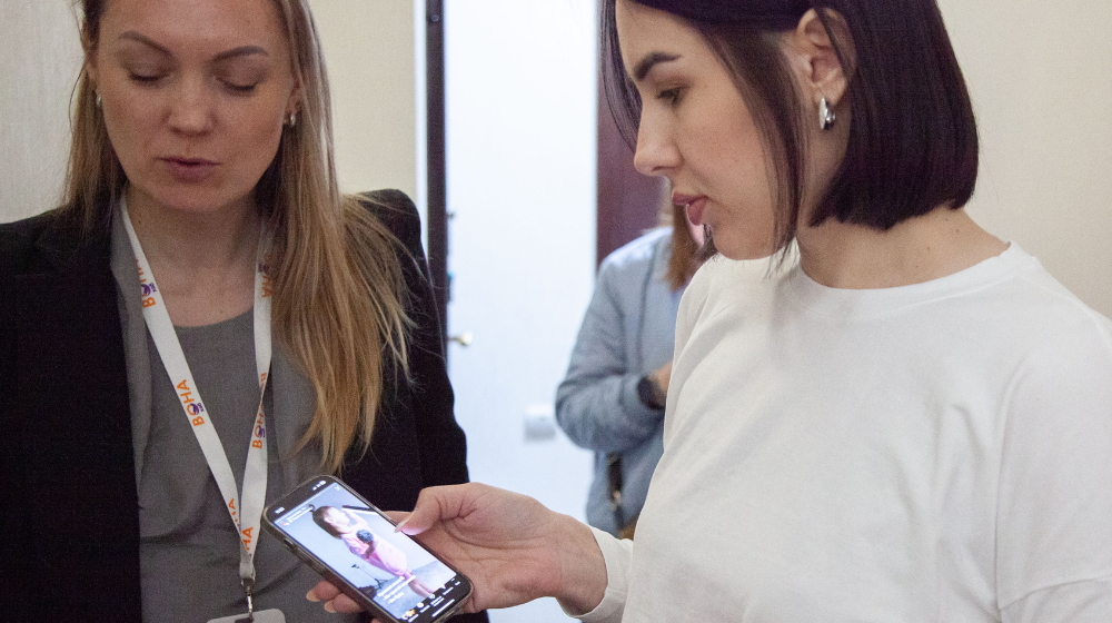 Woman in white shirt looks at a photo of children's clothing on her phone while standing next to woman in black blazer