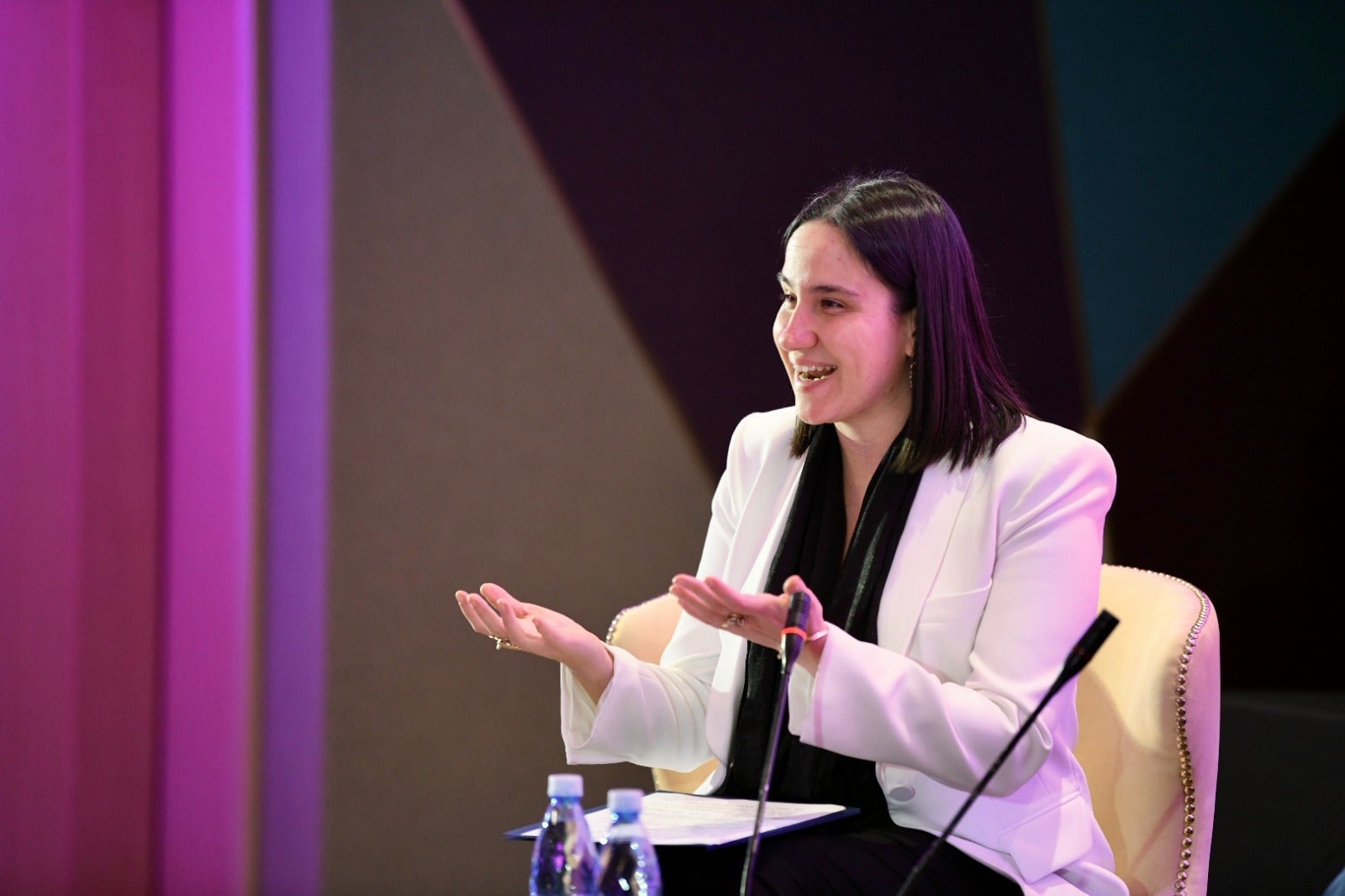 A woman in a black shirt and white blazer speaks on a conference stage